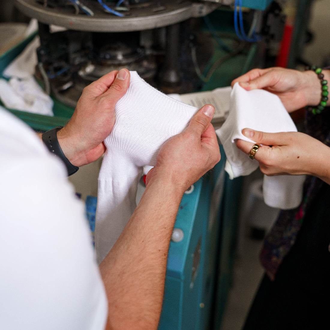 A close-up of a person's hands holding up a white knit garment, with another person's hands holding a matching white piece, in front of a teal and gray knitting machine.